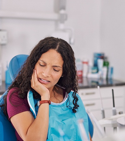 Woman in maroon shirt in dental chair with eyes closed holding hand to jaw